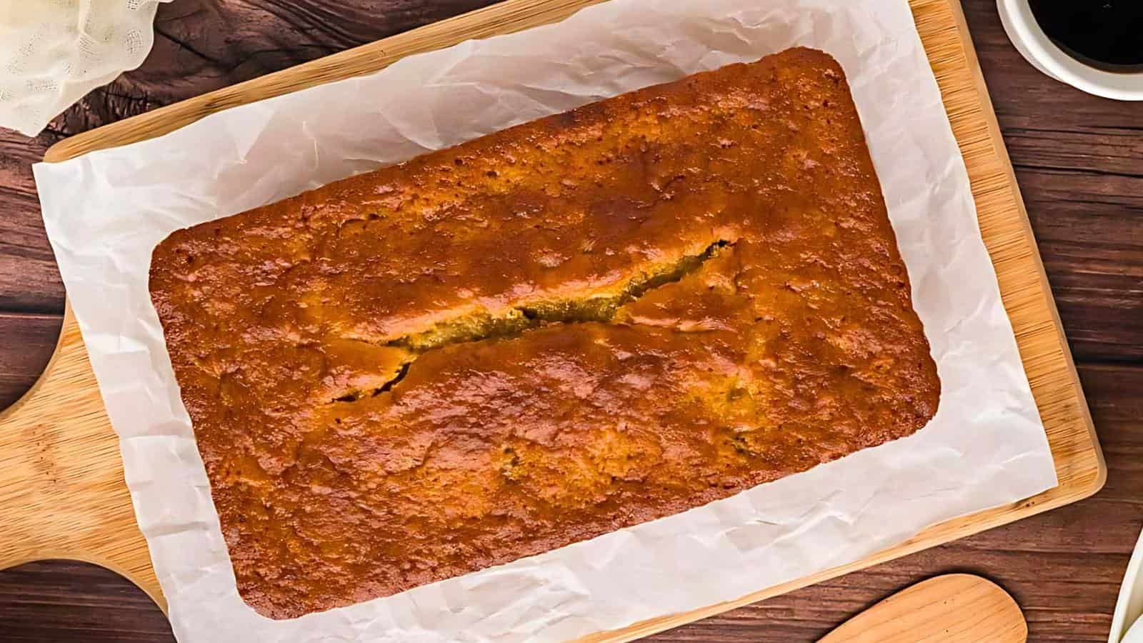 A freshly baked loaf of banana bread on a rectangular wooden cutting board lined with white parchment paper. The loaf has a golden-brown crust and a center crack. Part of a coffee cup and wooden spoon are visible in the corners of the image.
