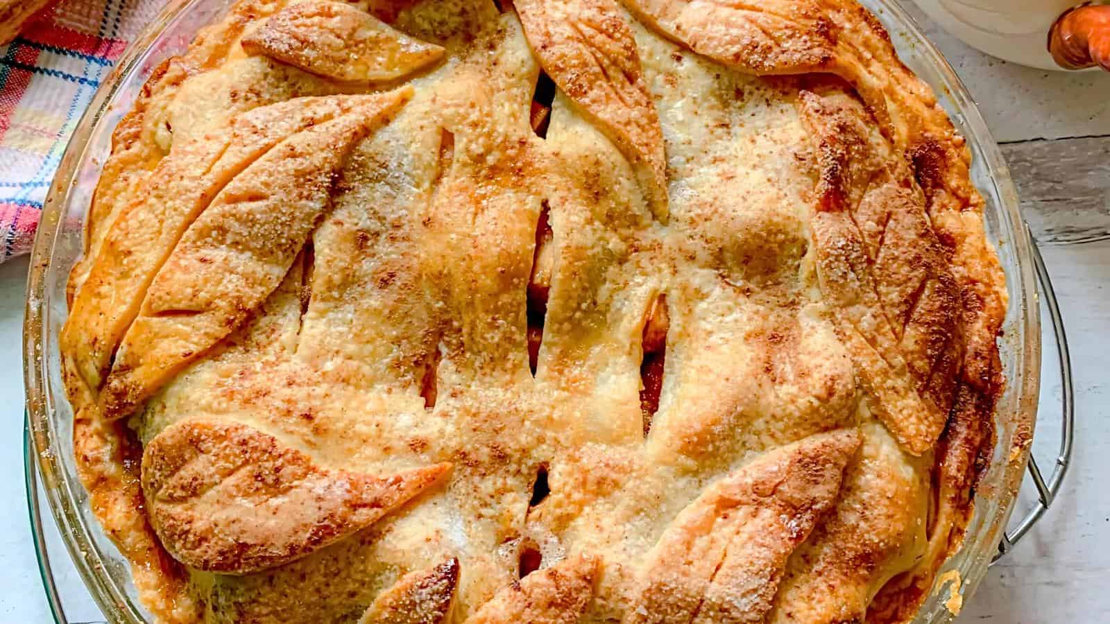 A golden-brown homemade pie topped with a flaky crust adorned with decorative leaf-shaped pastry cutouts. The crust is sprinkled with sugar, and diagonal slits reveal a glimpse of the filling inside. The pie is in a glass dish, ready for serving.