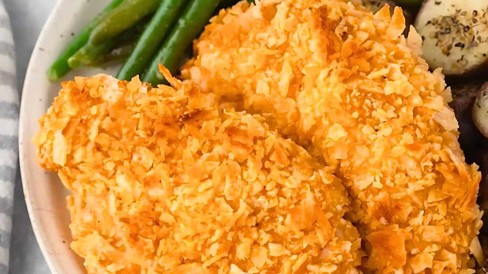 A close-up of crispy baked chicken tenders, golden and crunchy, served on a white plate with a side of green beans and roasted potatoes. The chicken tenders have a textured, breaded coating, creating an appetizing and inviting dish.