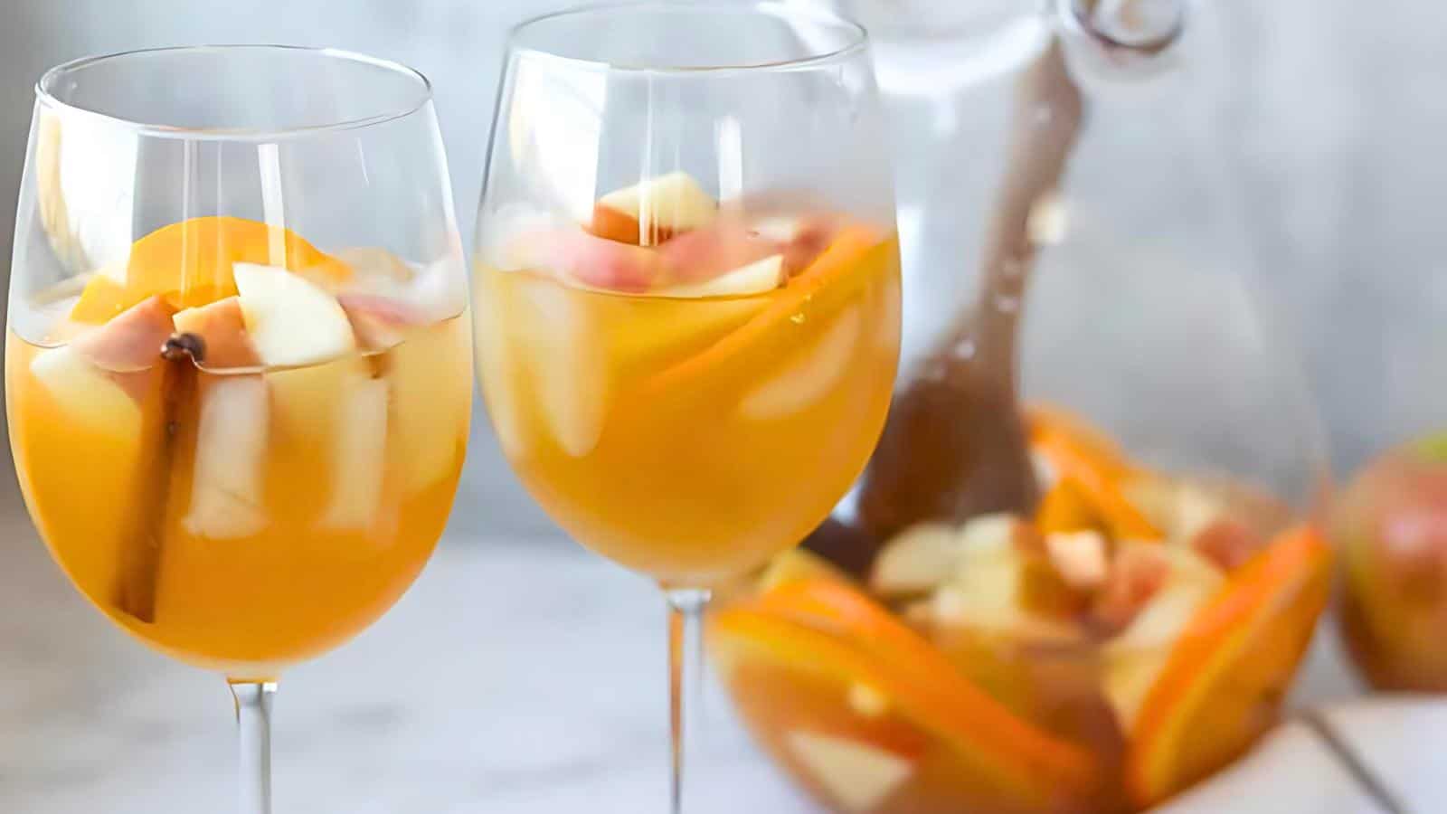 Two wine glasses filled with a refreshing fruit drink, featuring slices of apples, oranges, and a cinnamon stick. A pitcher containing more of the drink is visible in the background, set against a blurred white background.