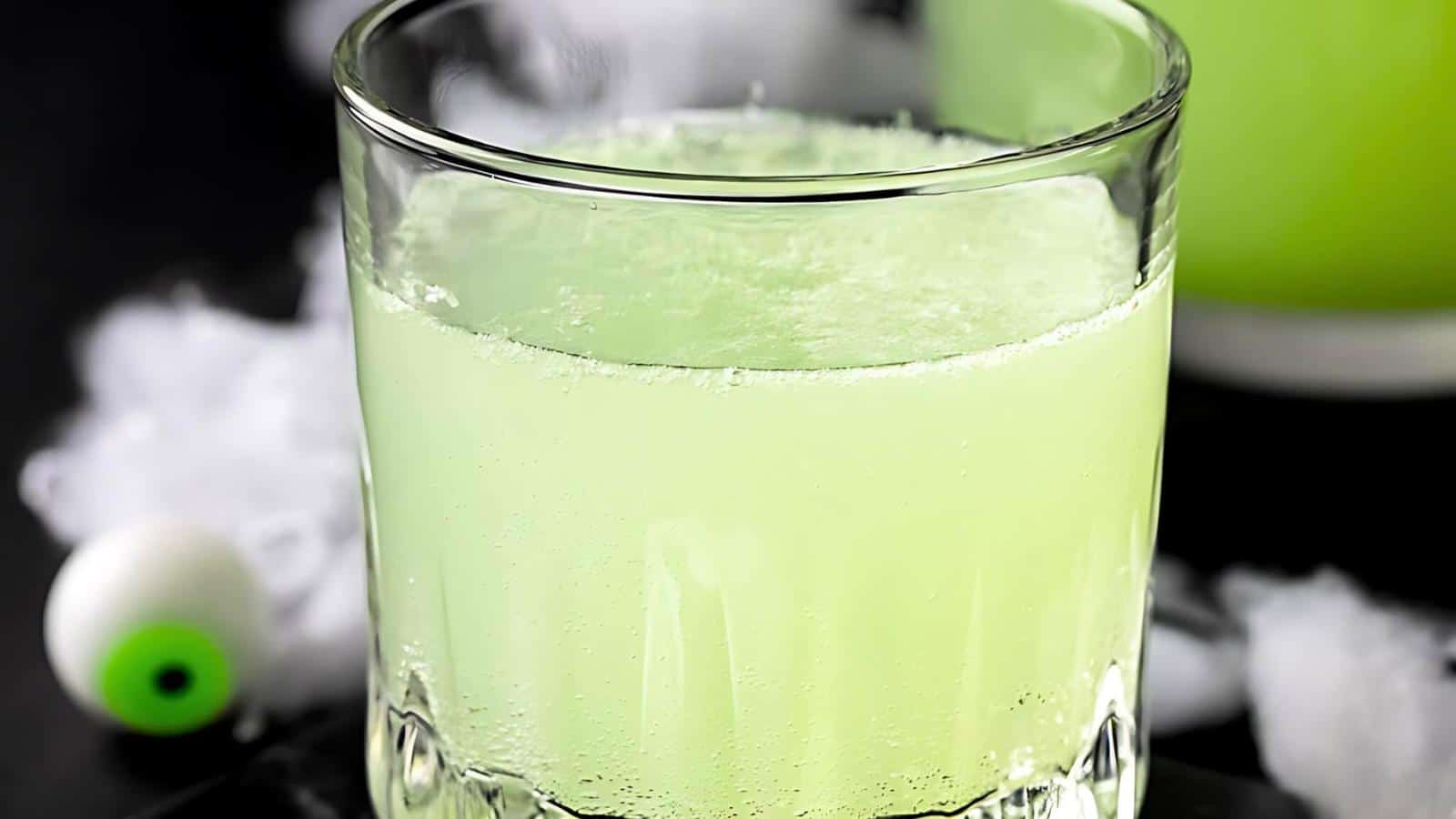 A clear glass filled with a bright green beverage, placed on a dark surface. In the background, there's a decor item resembling a white eyeball with a green iris, along with some white fog or smoke, creating a spooky ambiance.