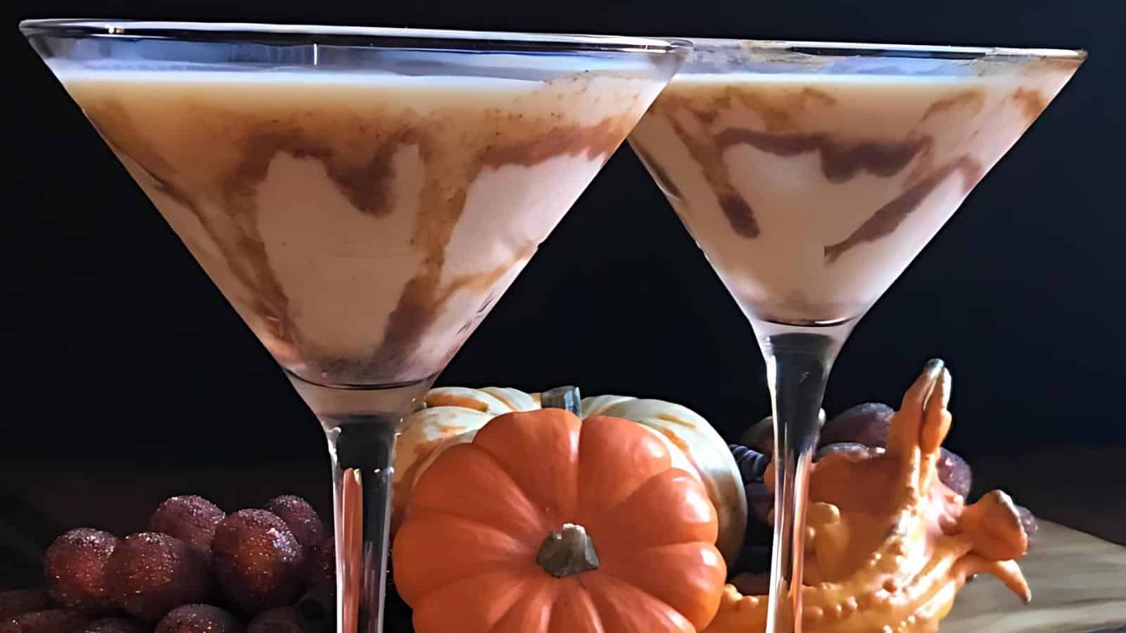 Two martini glasses filled with a creamy pumpkin spice cocktail are garnished with swirls of caramel. They are set amongst autumn decorations, including mini pumpkins, glittery acorns, and a decorative ceramic turkey, against a dark background.