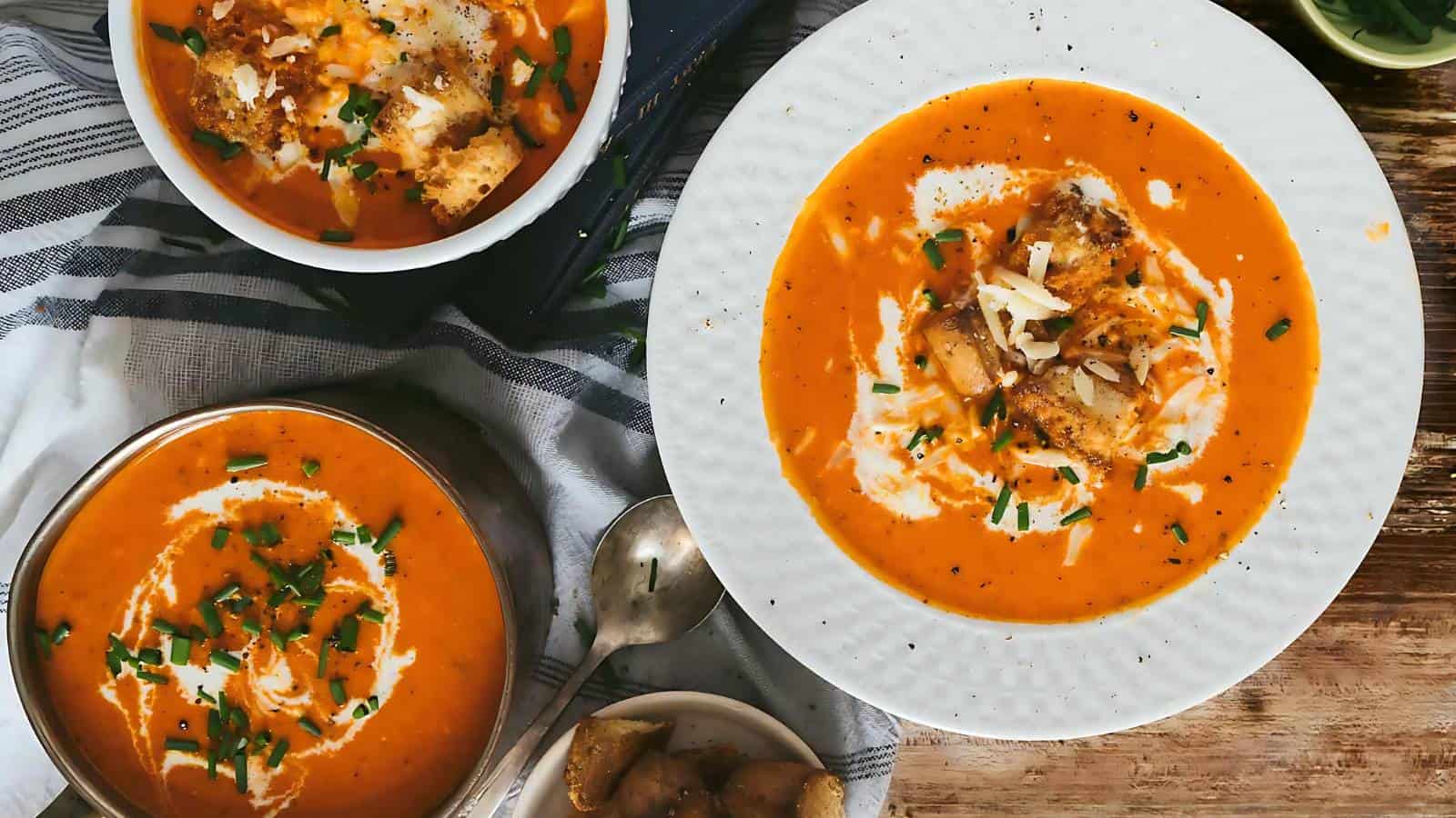 Three bowls of creamy tomato soup garnished with herbs and croutons are displayed on a table. One large bowl is placed on a white plate, and two smaller bowls are adjacent. A napkin and a pair of spoons are nearby, with a small dish of diced potatoes beside them.