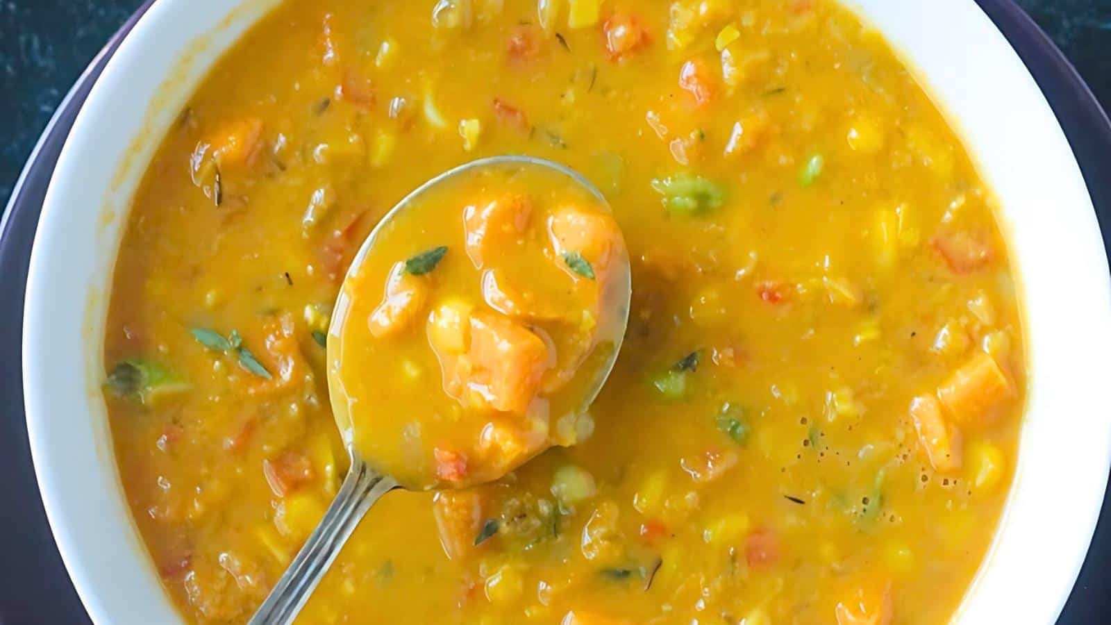 A bowl of hearty pumpkin soup filled with chunks of vegetables such as carrots, corn, and potatoes, garnished with fresh herbs. A spoon is scooping up a portion of the thick, golden-orange soup, showcasing the vibrant ingredients and rich texture.