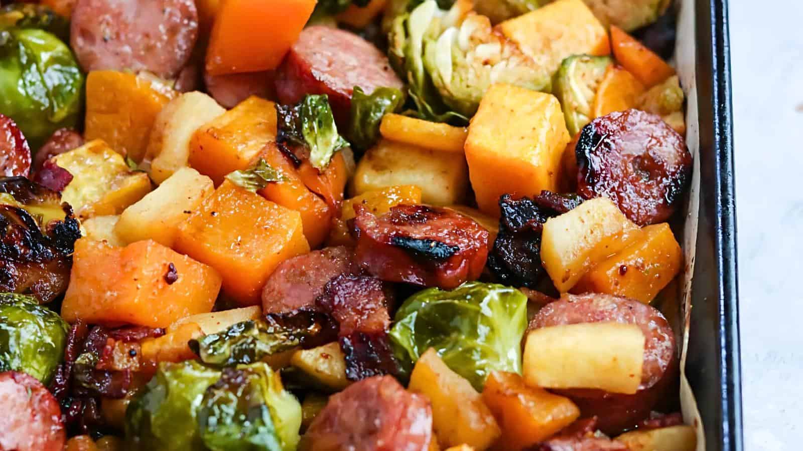 A close-up of a baked dish featuring roasted Brussels sprouts, chunks of sausage, sweet potatoes, and other root vegetables. The ingredients are caramelized and browned, suggesting a savory and hearty meal.
