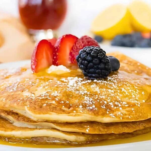 A stack of sourdough discard pancakes topped with sliced strawberries, a blackberry, and syrup, sprinkled with powdered sugar.