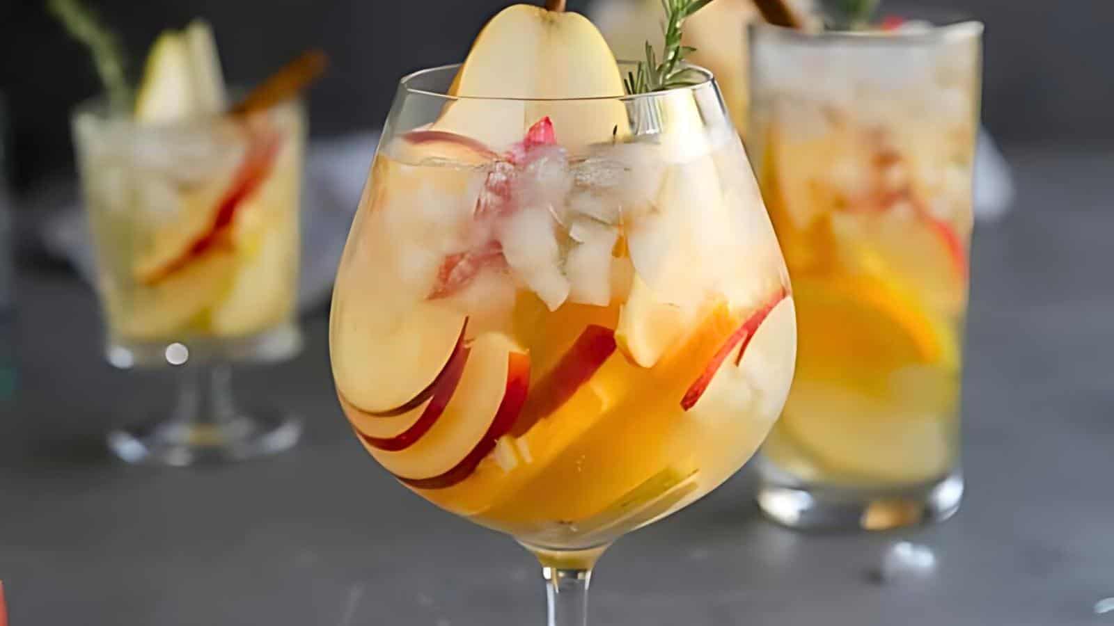 A close-up of a cocktail glass filled with a refreshing drink that includes ice cubes, apple slices, and orange slices. In the background, there are three similar glasses of the same drink, garnished with apple slices and herbs.
