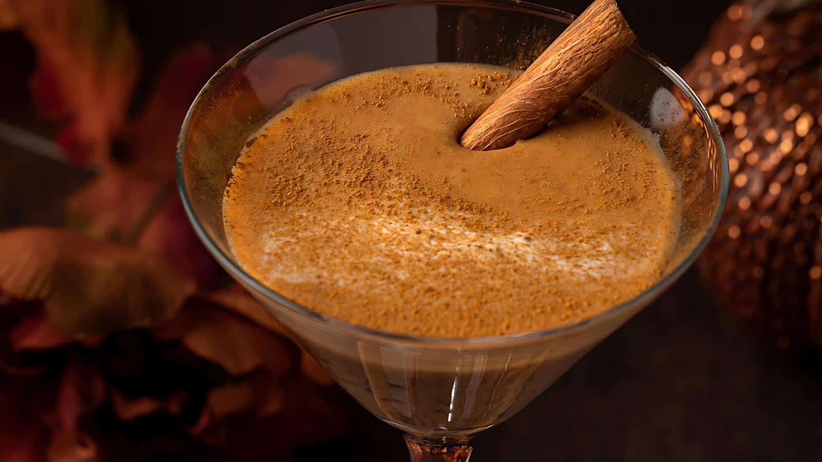 A close-up view of a creamy brown cocktail in a martini glass, garnished with a cinnamon stick and a dusting of ground cinnamon on top. The background features warm, out-of-focus autumnal decor, enhancing the cozy and festive atmosphere.