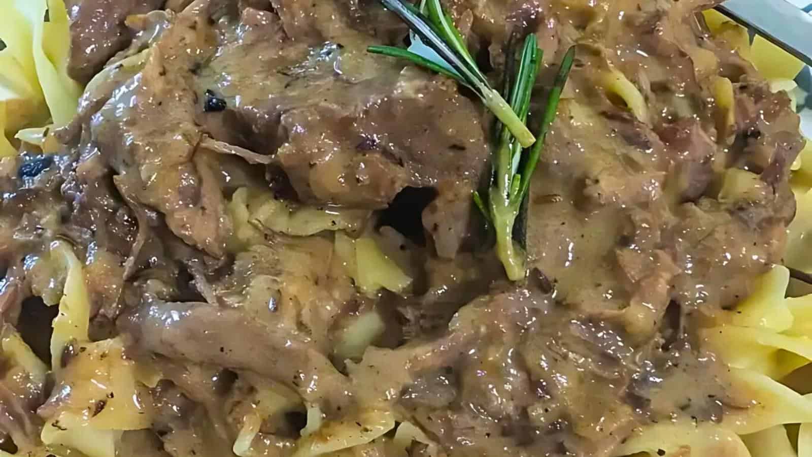 Close-up of a creamy pasta dish topped with shredded meat and garnished with sprigs of fresh rosemary. The pasta is coated in a thick sauce, rich in texture, and the meat appears tender and succulent.