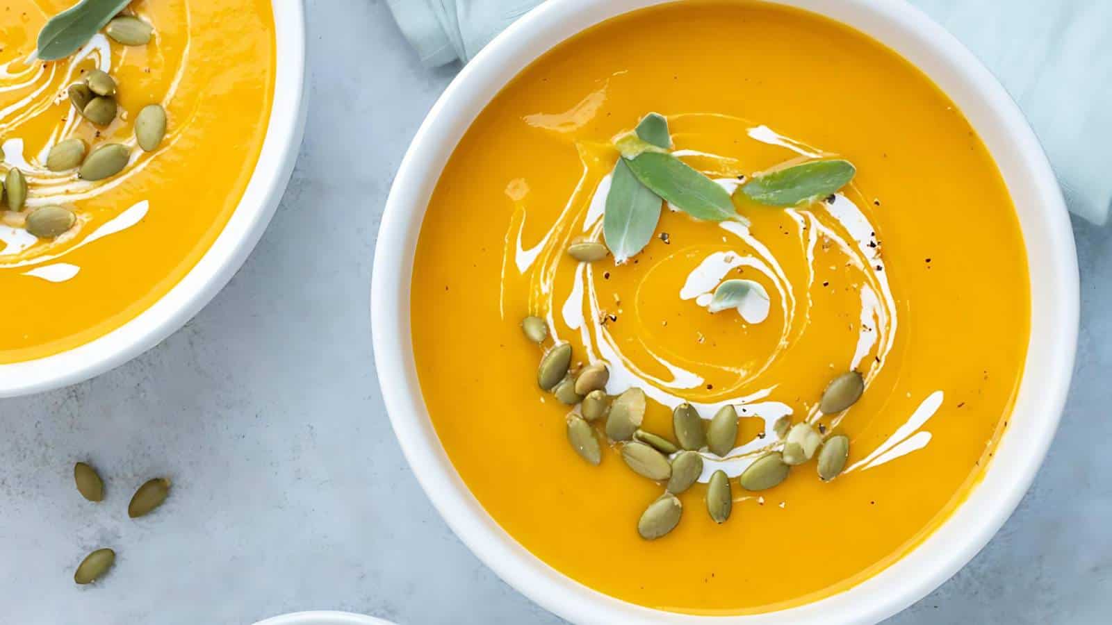 A bowl of vibrant orange soup garnished with a swirl of cream, pumpkin seeds, and fresh sage leaves. The bowl is placed on a light-colored surface, with another identical bowl and scattered pumpkin seeds visible in the background.