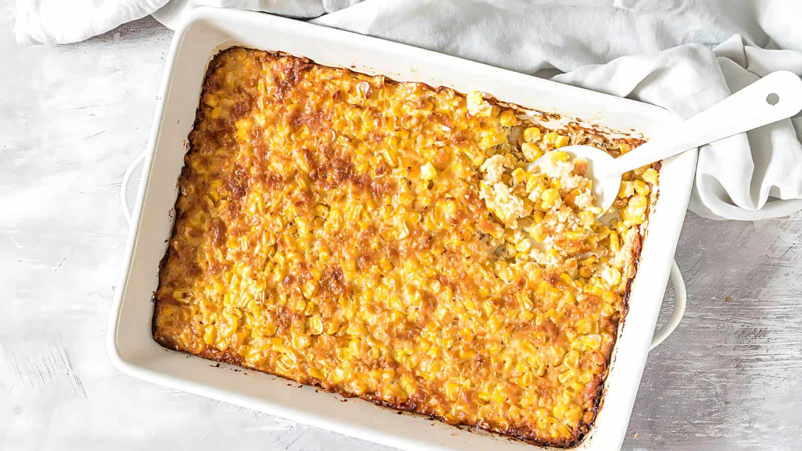 A baked corn casserole in a white rectangular dish, partially scooped out with a white serving spoon. The casserole has a golden-brown crust and a creamy texture, sitting on a white surface with a light-colored cloth in the background.