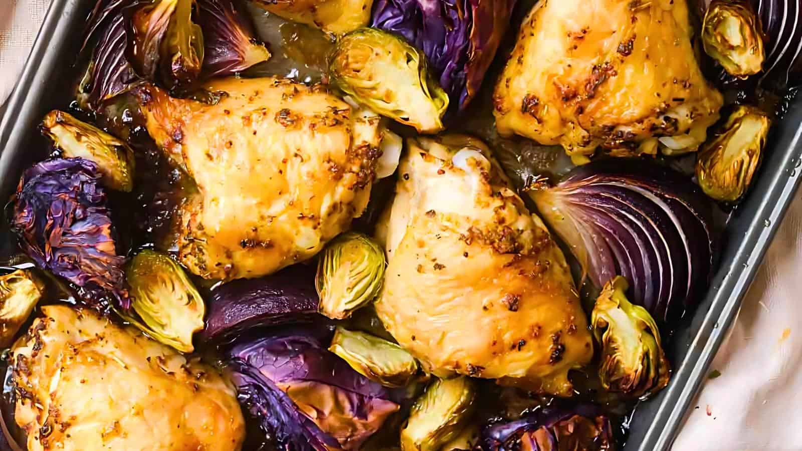A close-up of a roasted chicken dish in a baking pan. The chicken thighs are golden-brown and seasoned, surrounded by browned Brussels sprouts and red cabbage wedges. The dish appears to be freshly baked and glistening with a light sauce.
