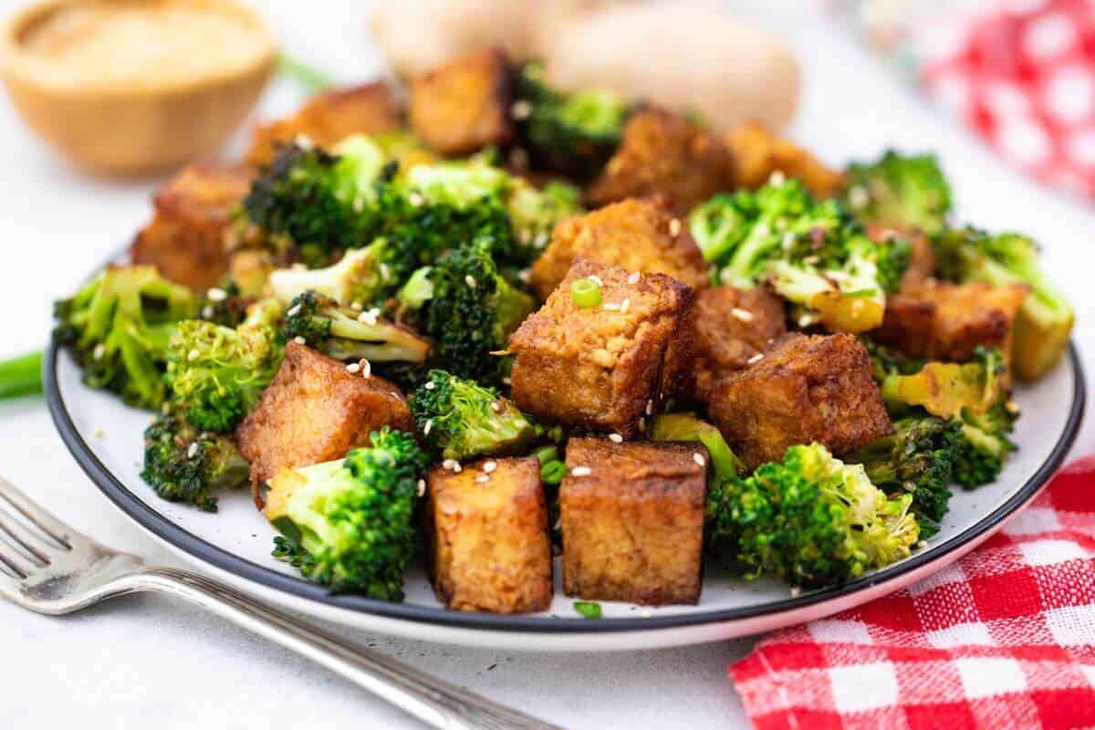 A plate of stir-fried tofu and broccoli garnished with sesame seeds is shown. The tofu is golden brown and cubed, nestled among bright green broccoli florets. A fork and a red checkered cloth napkin are positioned nearby.