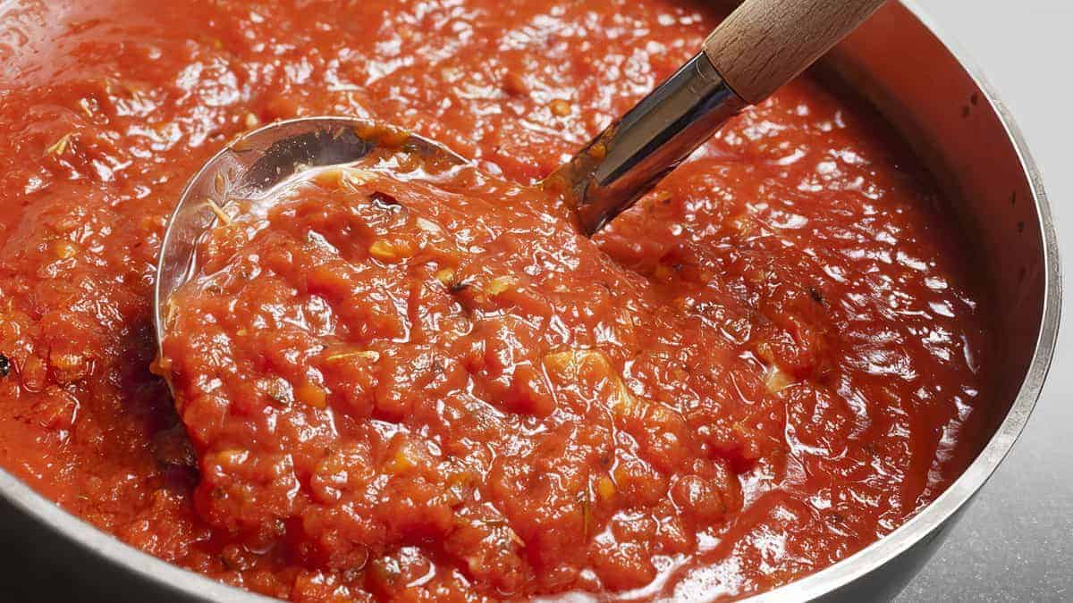 A close-up of a ladle scooping a hearty, chunky tomato sauce from a large stainless steel pot. The sauce has a rich red color and visible pieces of tomatoes and herbs, indicating a thick and flavorful consistency. The ladle has a wooden handle.