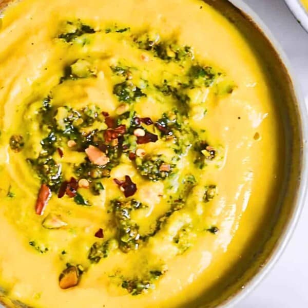 A bowl of yellow soup garnished with green herbs and red chili flakes on a white surface.
