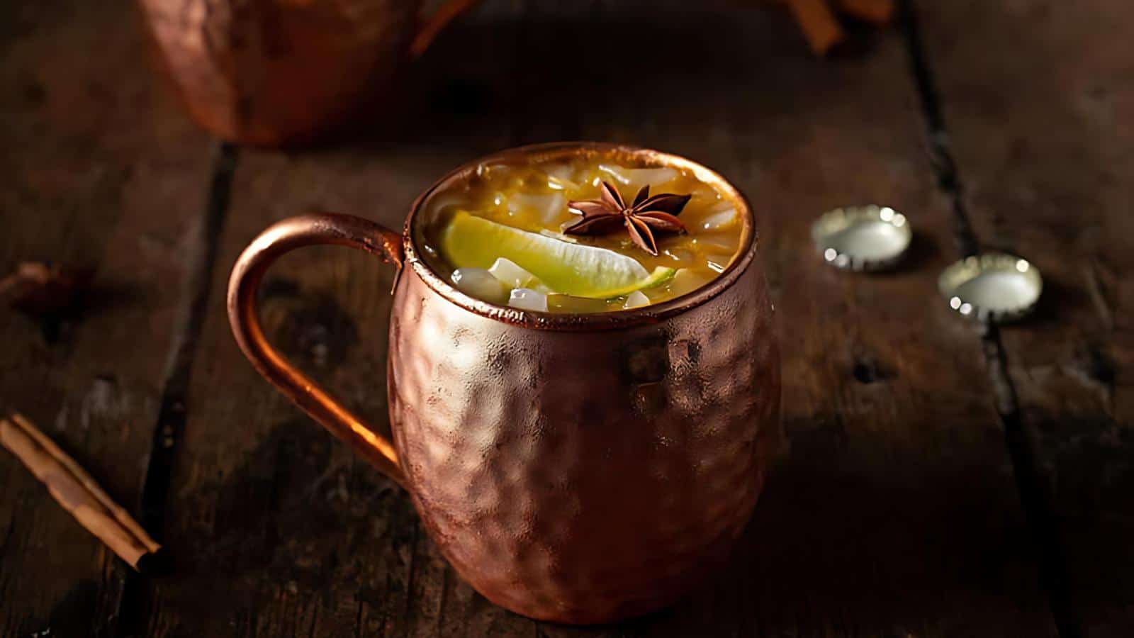 A copper mug filled with a spiced cocktail garnished with lemon slices and star anise sits on a rustic wooden table. In the background, bottle caps and part of another copper cup are visible.