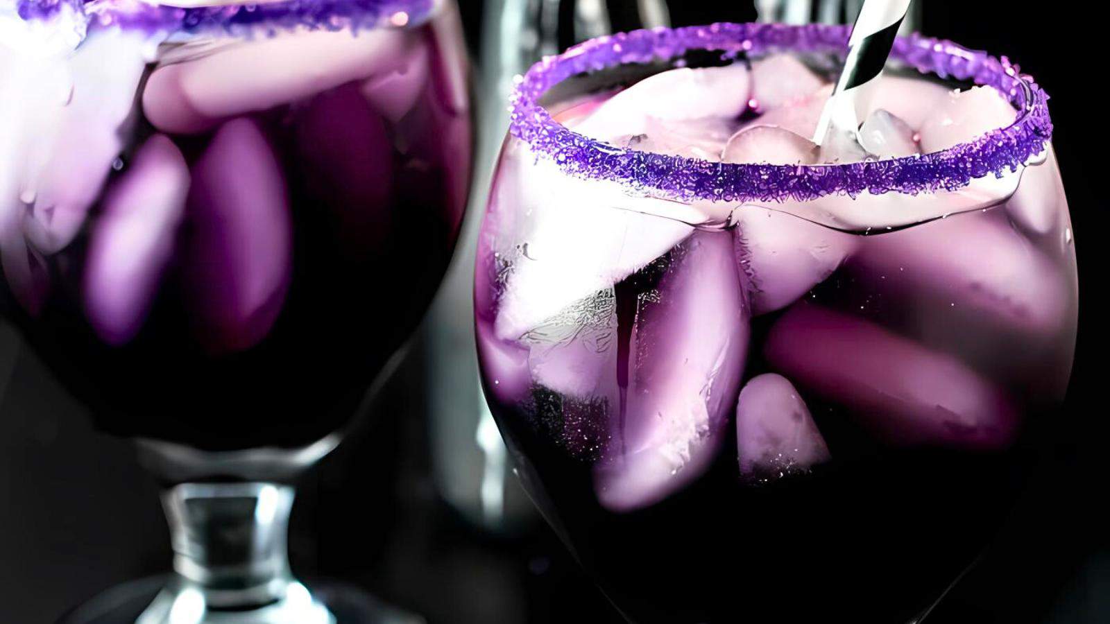 Close-up of two stemmed glasses filled with vibrant purple beverages and ice cubes. The rims are coated with purple sugar crystals, and a black and white striped straw is in one of the glasses. The background is dark, making the drinks stand out.