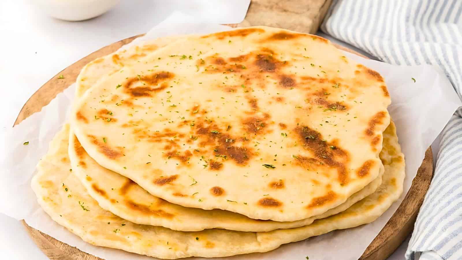 A stack of golden brown naan breads sprinkled with herbs, placed on a piece of parchment paper on a wooden board. In the background, there is a striped cloth and part of a white bowl.