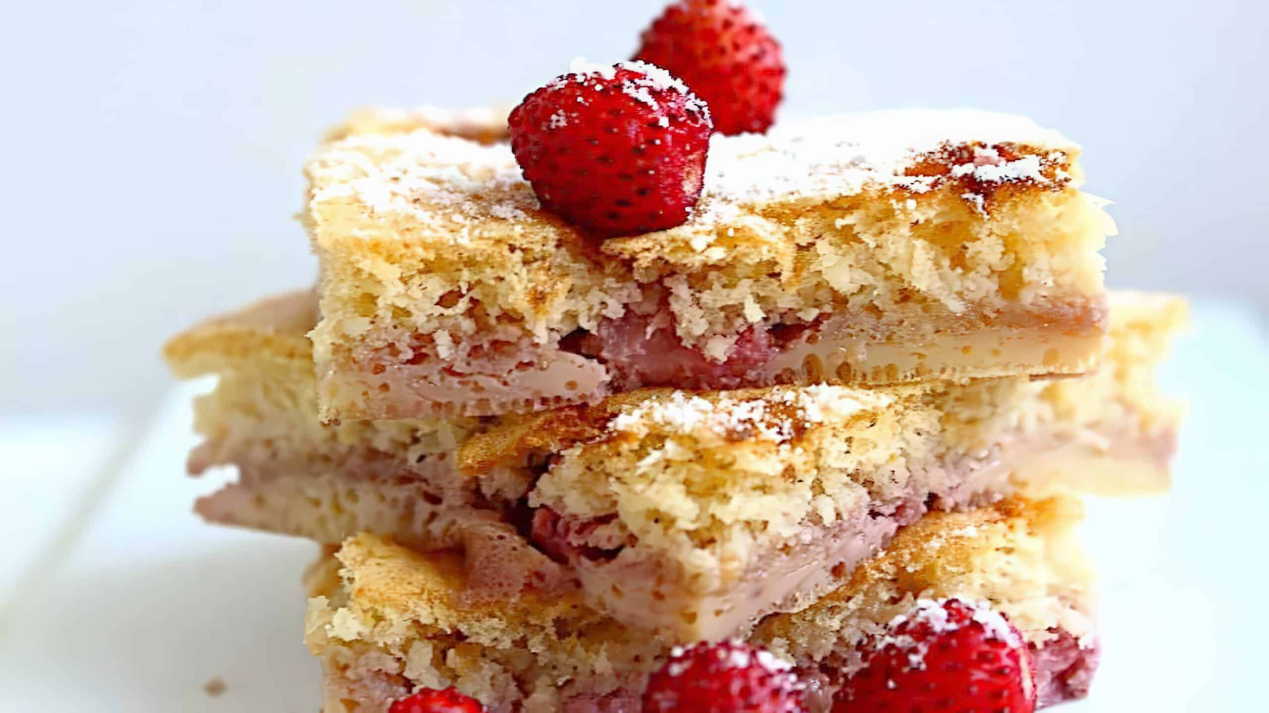 A stack of three strawberry shortcake squares are topped with whole strawberries and a dusting of powdered sugar. The shortcake looks soft and fluffy, with visible layers of strawberry filling in between the cake layers.
