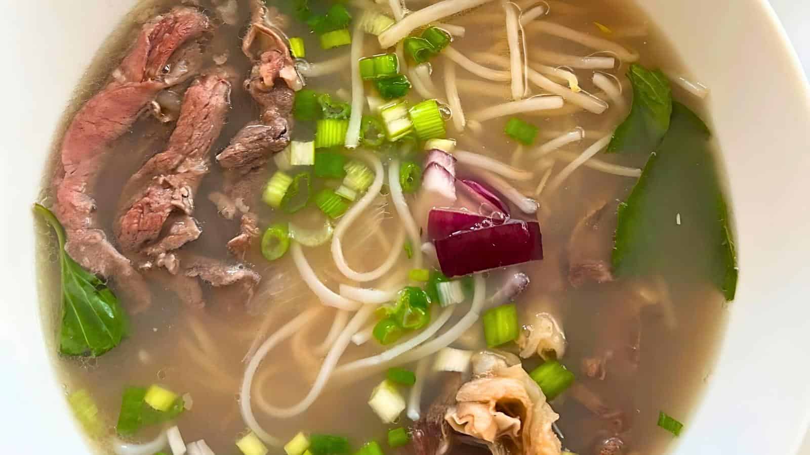 A bowl of Vietnamese pho soup is filled with thin rice noodles, sliced rare beef, red onions, green onions, and fresh herbs. The clear broth looks flavorful and steamy, with pieces of greens floating on the surface.