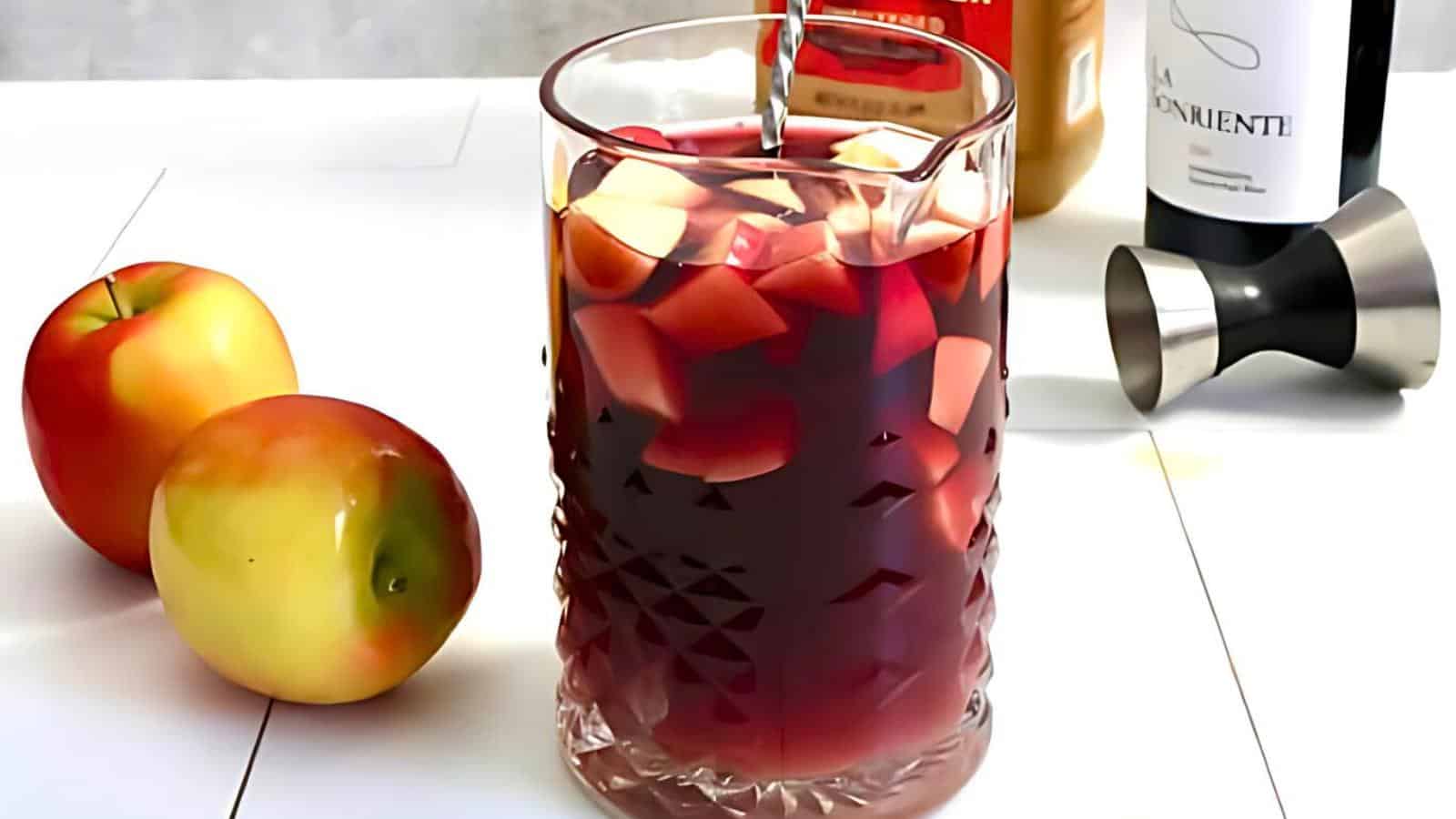 A glass of sangria filled with chopped fruit sits on a white surface. Next to the glass are two apples, a jigger, and bottles of wine and juice in the background. The sangria has a rich, deep red color, and the fruit pieces are visible through the glass.