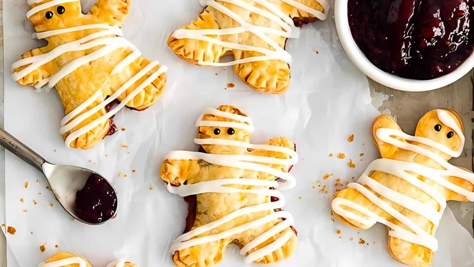 A collection of mummy-shaped pastries, drizzled with white icing to resemble bandages, placed on a white parchment paper. A spoon with dark jam and a bowl filled with jam are also visible beside the pastries.