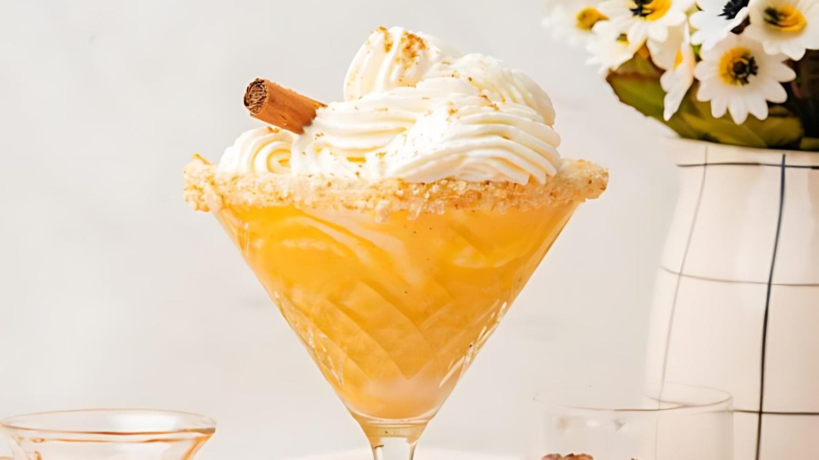 A festive cocktail in a martini glass, topped with swirls of whipped cream and garnished with a cinnamon stick, sits on a white surface. The rim is coated with sugar, and a vase of white flowers is in the background, slightly blurred.
