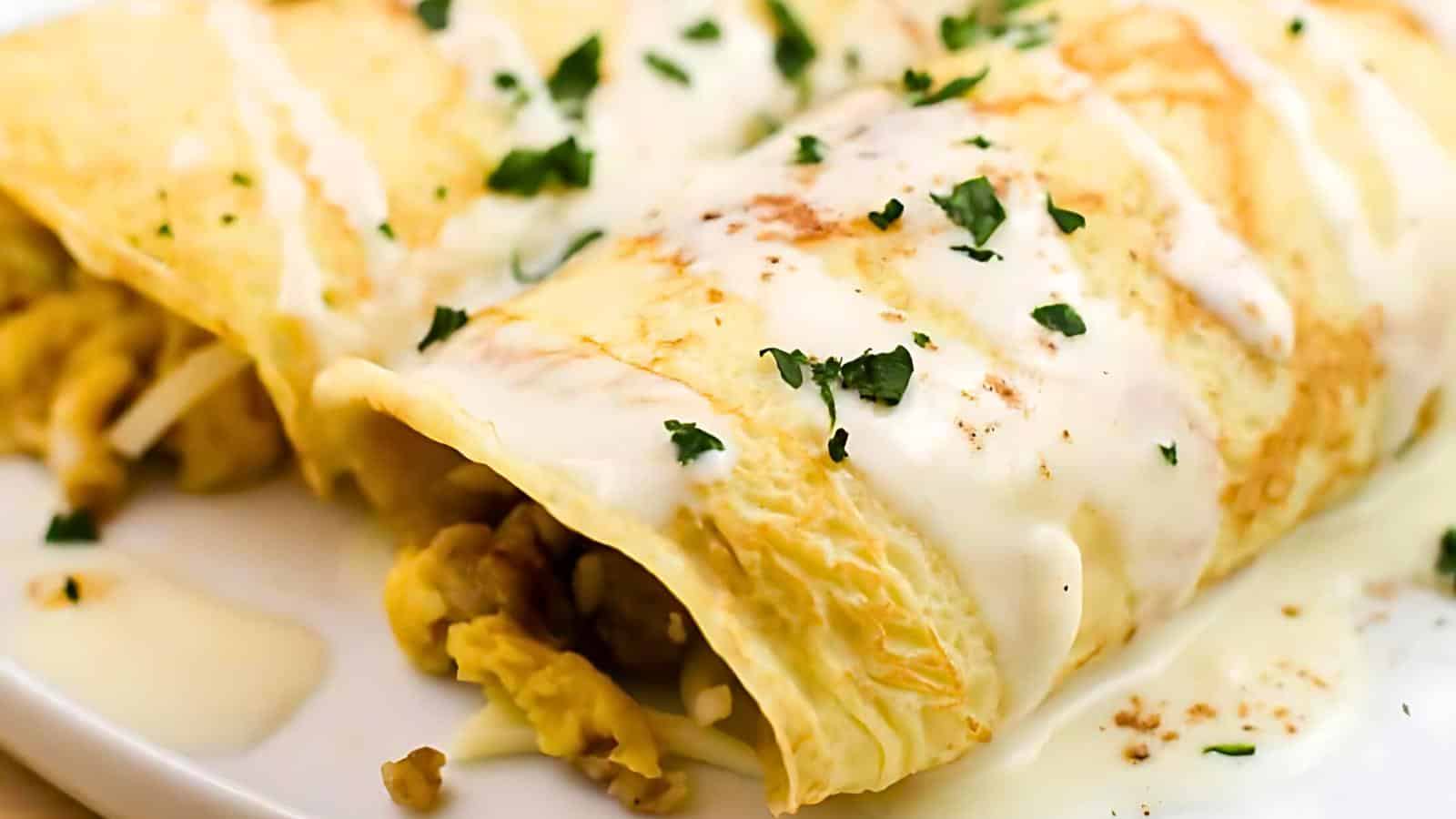 A close-up of a cheesy breakfast omelet filled with scrambled eggs and vegetables, topped with drizzles of creamy white sauce and sprinkled with fresh chopped parsley. The omelet sits on a white plate.