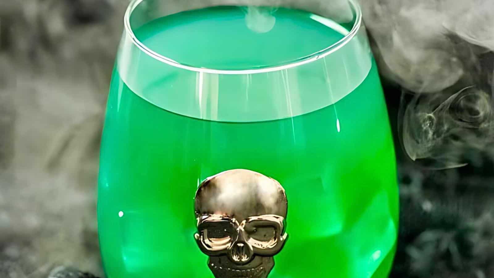 A green drink served in a rounded glass is adorned with a metallic skull ornament. Wisps of vapor emanate around the glass, creating a misty effect. The spooky presentation evokes a sense of mystery and intrigue.