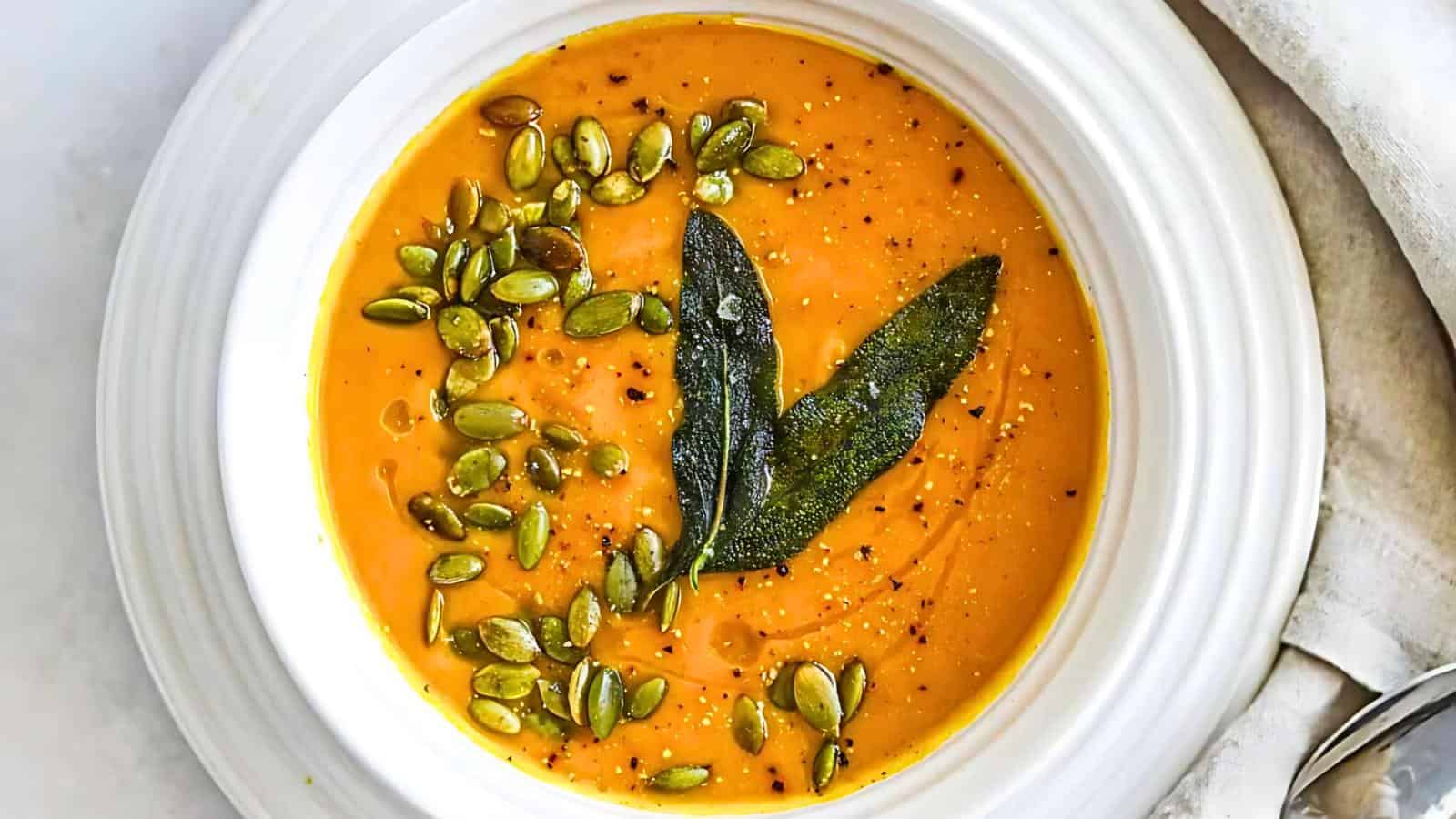 A bowl of creamy orange soup garnished with roasted pumpkin seeds and two sage leaves. The soup is served in a white bowl on a white surface, with a beige napkin and a spoon partially visible to the side.