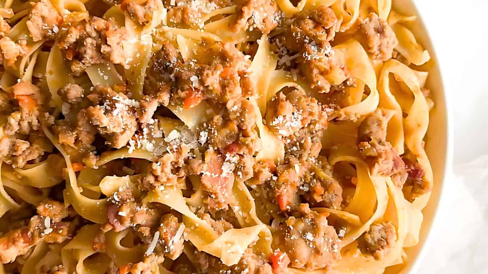 A close-up of a plate of tagliatelle pasta topped with a rich meat sauce, garnished with grated Parmesan cheese. The sauce appears to have ground meat and finely chopped vegetables, creating a hearty and savory dish.
