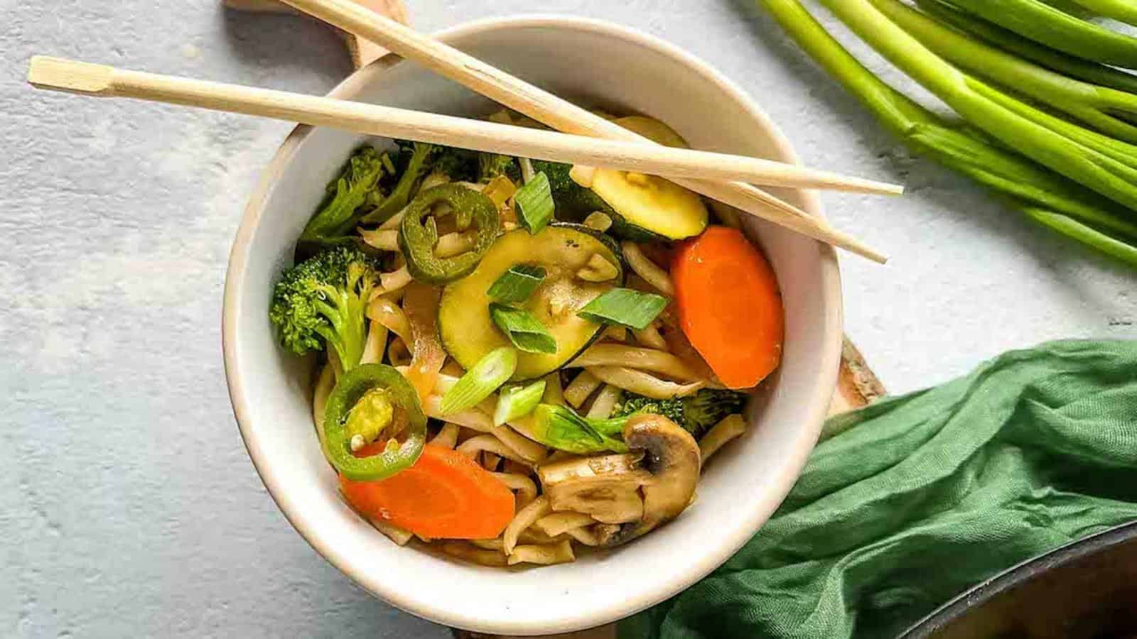 A bowl of stir-fried noodles with sliced vegetables including broccoli, carrots, mushrooms, and zucchini, garnished with sliced green onions and jalapeños. The bowl is accompanied by a pair of wooden chopsticks and a green cloth, with green onions visible in the background.
