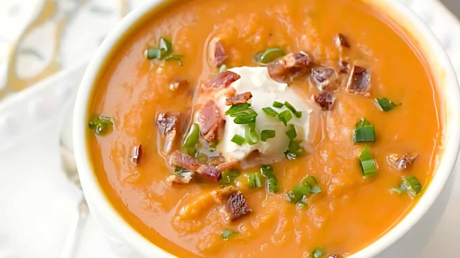 A close-up of a bowl of creamy orange soup, garnished with a dollop of sour cream, chopped bacon bits, and fresh chopped green onions. The bowl is set on a white plate, and a spoon is partially visible in the background.