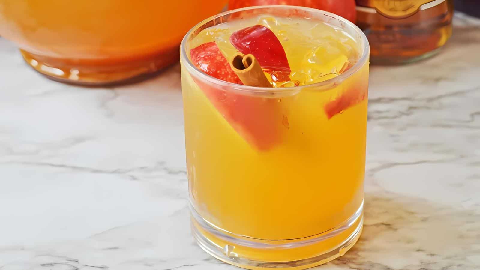 A glass filled with orange-colored liquid, garnished with red apple slices and a cinnamon stick, placed on a marble surface. In the background, there is a glass pitcher with more of the same orange beverage and a bottle with a bronze cap.