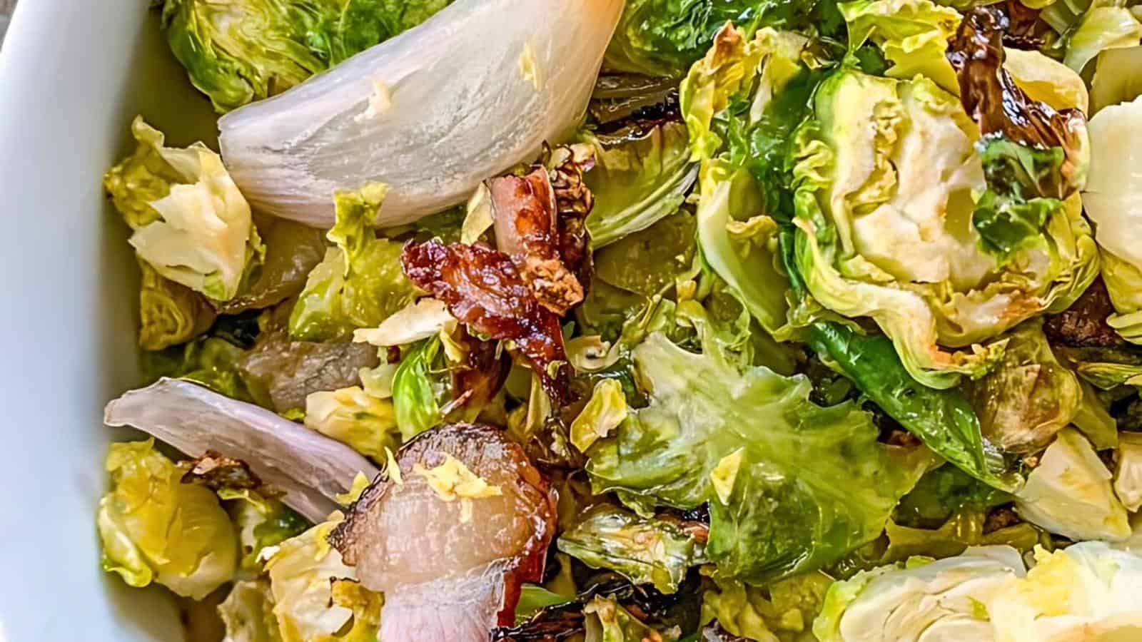 A close-up image of a bowl filled with roasted Brussels sprouts, caramelized onions, and crispy bacon pieces. The vegetables are charred in spots, giving them a crispy appearance, and the dish appears to be well-seasoned.