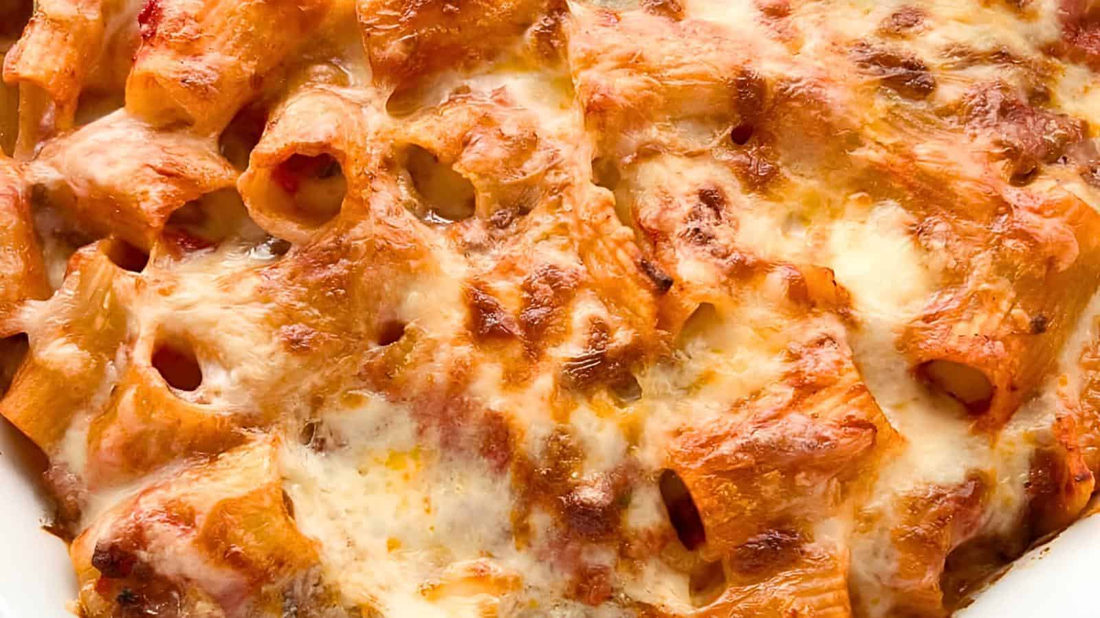 Close-up of baked ziti with melted cheese on top. The dish features tube-shaped pasta covered in a golden-brown layer of melted mozzarella and parmesan cheese, with visible spots of sauce and browned, crispy edges.