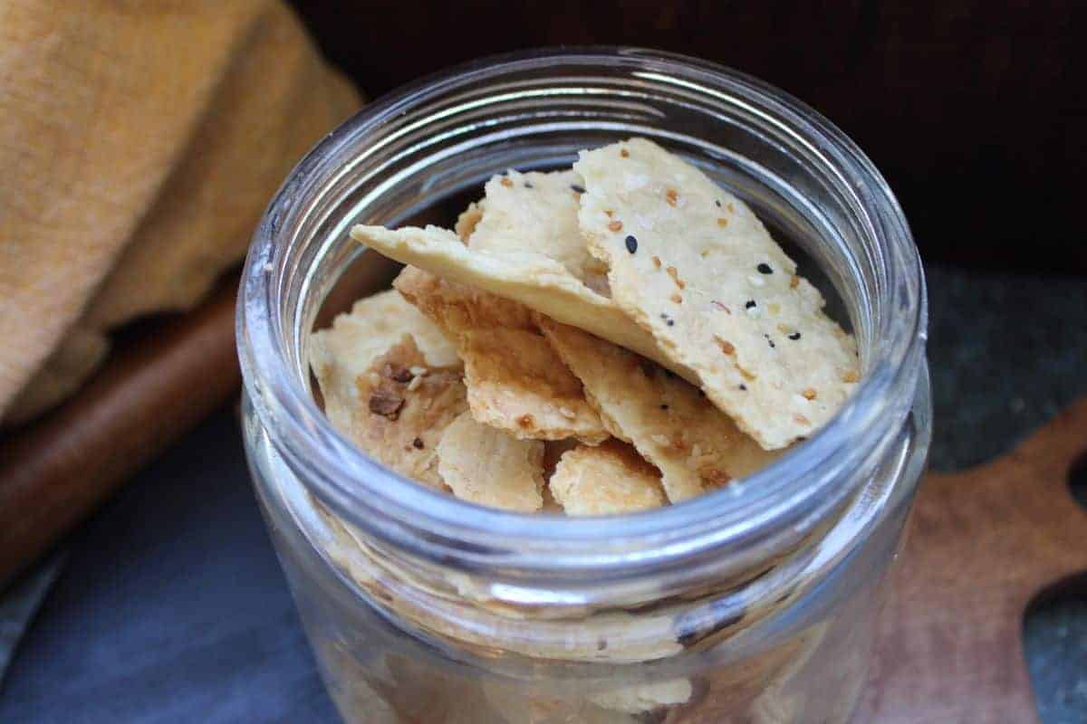 A close-up view of a glass jar filled with square-shaped, herb-dusted crackers. The jar is filled to the brim and the crackers have visible seasoning including seeds and spices. The background includes a rustic cloth and wooden surface.