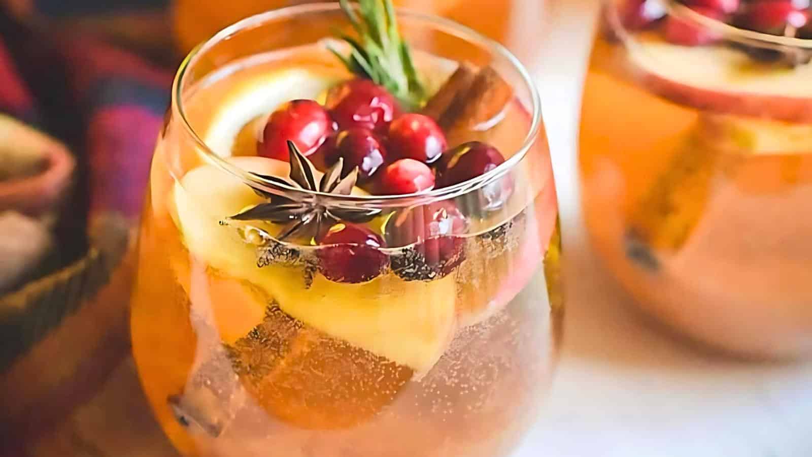 A close-up of a glass containing a festive holiday drink. Garnished with slices of apple, lemon, cinnamon sticks, star anise, cranberries, and a sprig of rosemary, the drink appears to be a sparkling beverage. Other similar glasses are visible in the background.