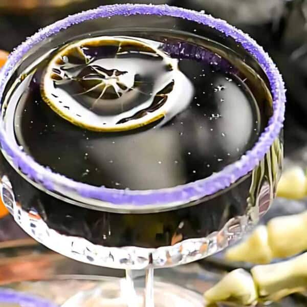 A black Halloween cocktail with a lemon wheel in a purple sugar-rimmed glass. A small pumpkin and skeleton hand are in the background.