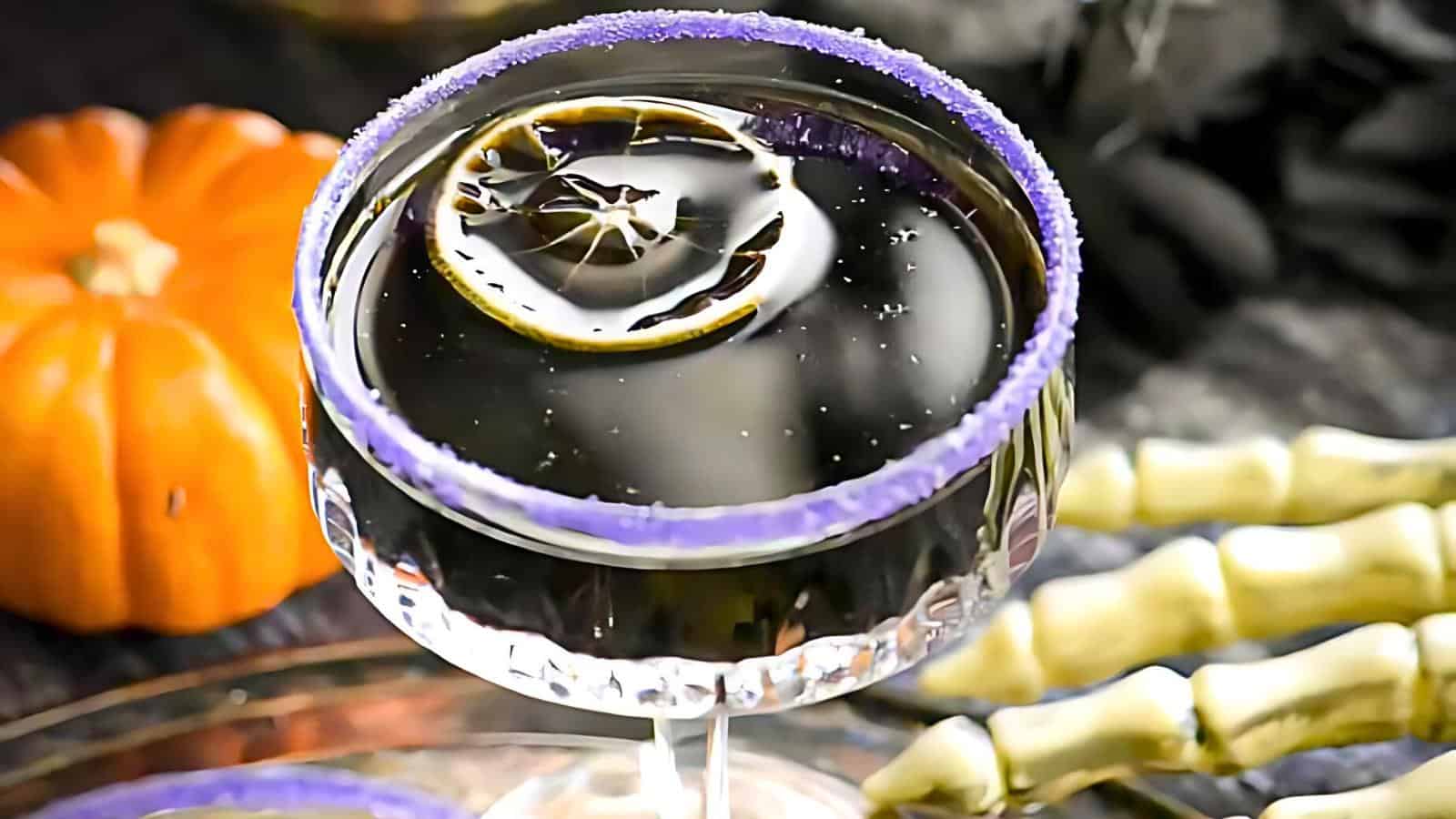 A spooky black cocktail in a glass with a purple sugar rim, garnished with a slice of citrus fruit. In the background, there is an orange pumpkin and skeleton hands, creating a Halloween-themed setting.