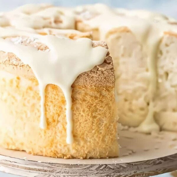 A creamy, white frosted cake with a slice removed, revealing a light and airy interior. The cake sits on a white stand against a light blue background.