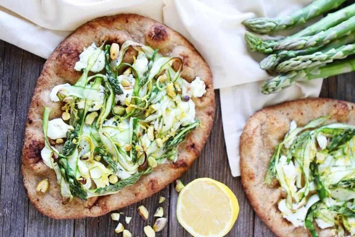 Two pieces of flatbread topped with ricotta cheese, shaved asparagus, and chopped pistachios are laid out on a wooden surface. A cut lemon and fresh asparagus spears are placed nearby on a white cloth. The scene conveys a fresh, appetizing meal.