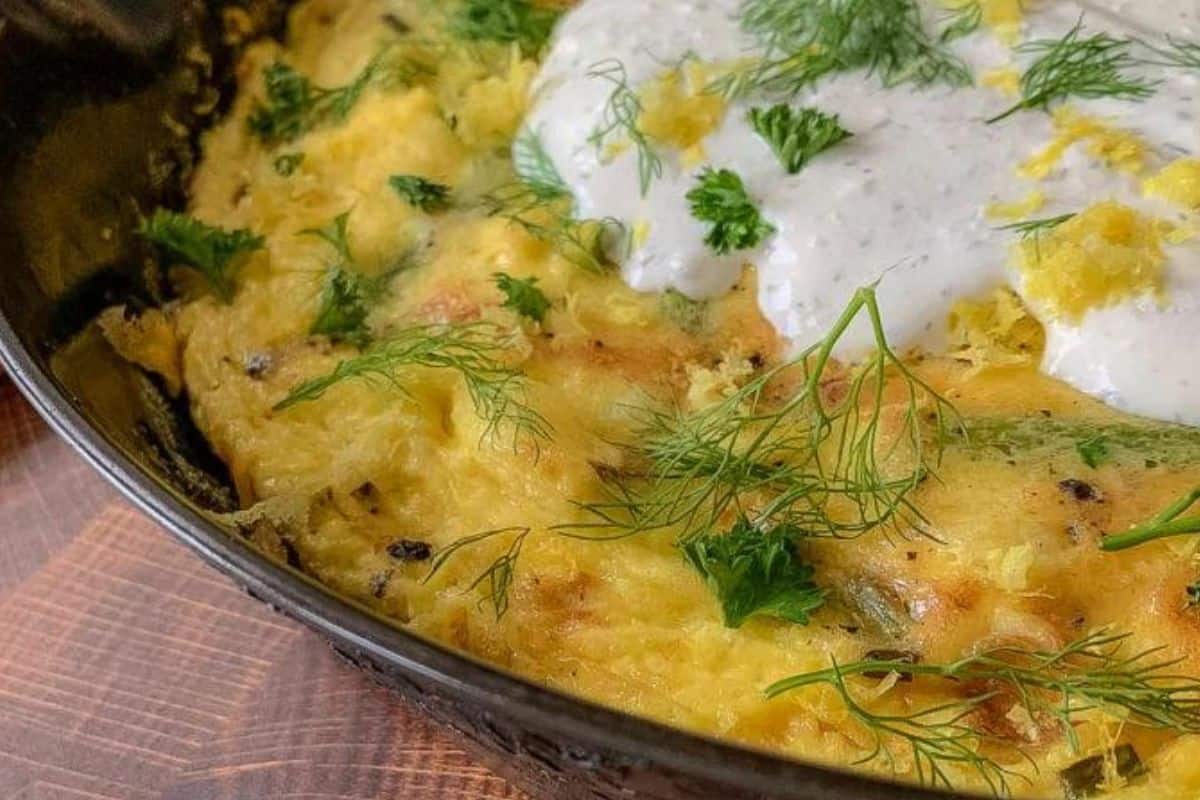 A close-up of a pan containing a golden-brown baked dish topped with a creamy white sauce and garnished with fresh herbs including dill and parsley. The dish is placed on a wooden surface.