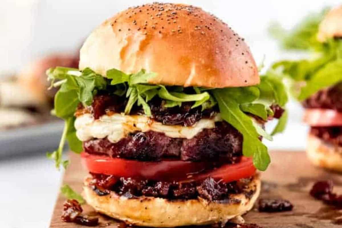 A close-up of a gourmet burger with a poppy seed bun, filled with fresh arugula, caramelized onions, creamy cheese, a thick beef patty, and a slice of tomato. The burger is placed on a wooden surface, with another similar burger slightly blurred in the background.