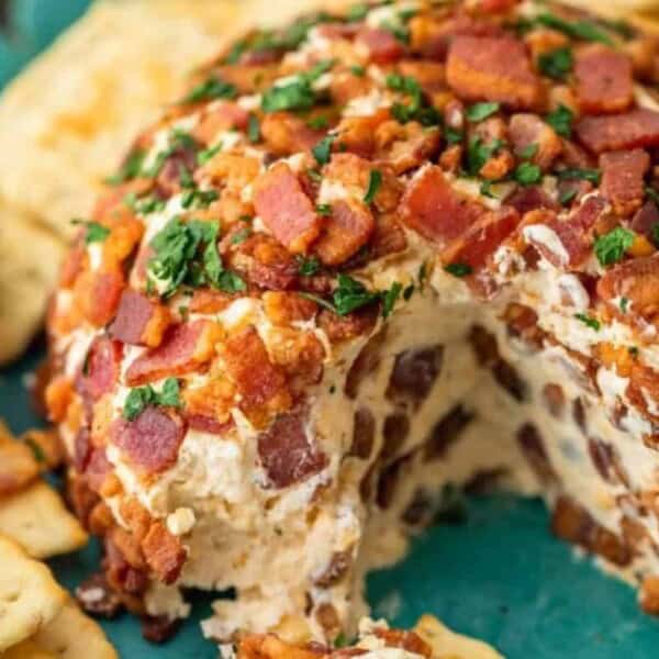 A cheese ball covered in chopped bacon and herbs sits on a plate. A portion is removed, revealing the creamy inside. Crackers and a spreading knife are placed nearby.