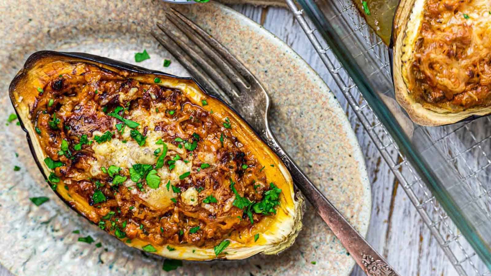 A baked stuffed eggplant sits on a plate, topped with a golden-brown mixture of minced vegetables and cheese, garnished with chopped fresh parsley. A fork rests beside the eggplant, and part of another stuffed eggplant is visible on a cooling rack.