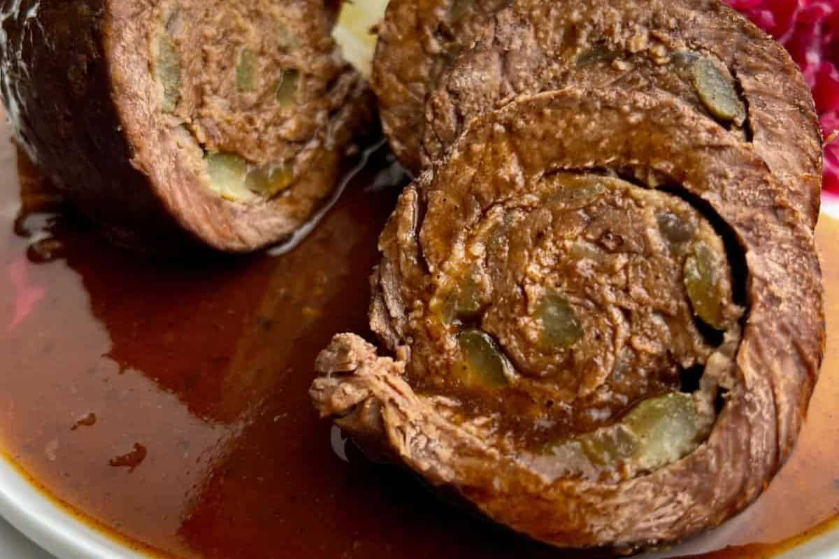 Close-up of sliced beef roulade on a white plate, filled with pickles and possibly other ingredients, with a rich brown gravy. The background shows some red cabbage on the right side. The meat appears tender and juicy, with a well-cooked, savory appearance.