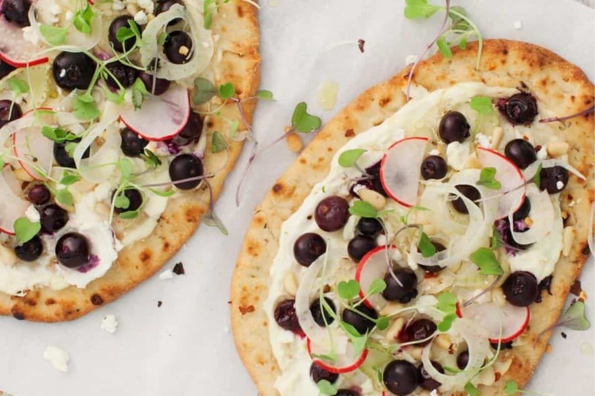 Two pieces of flatbread topped with a creamy spread, blueberries, radish slices, microgreens, and what appears to be crumbled cheese. The flatbreads are displayed on parchment paper.