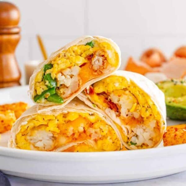 Three breakfast burritos stacked on a plate with tater tots and a halved avocado in the background.
