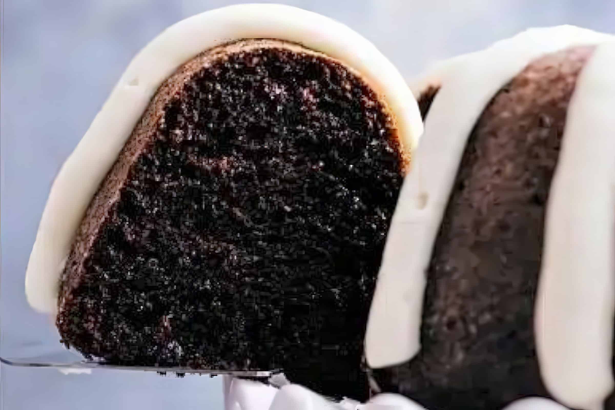 A close-up of a slice of chocolate bundt cake on a spatula. The cake is rich and moist, topped with white frosting that drips elegantly down the sides. The background is blurred, keeping the focus on the delicious slice.